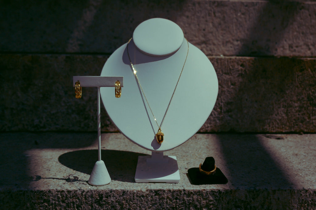 Gold dainty acorn necklace on white neck bust. Displayed alongside meander ring and earrings. On a granite rock background with creative purple lighting. 