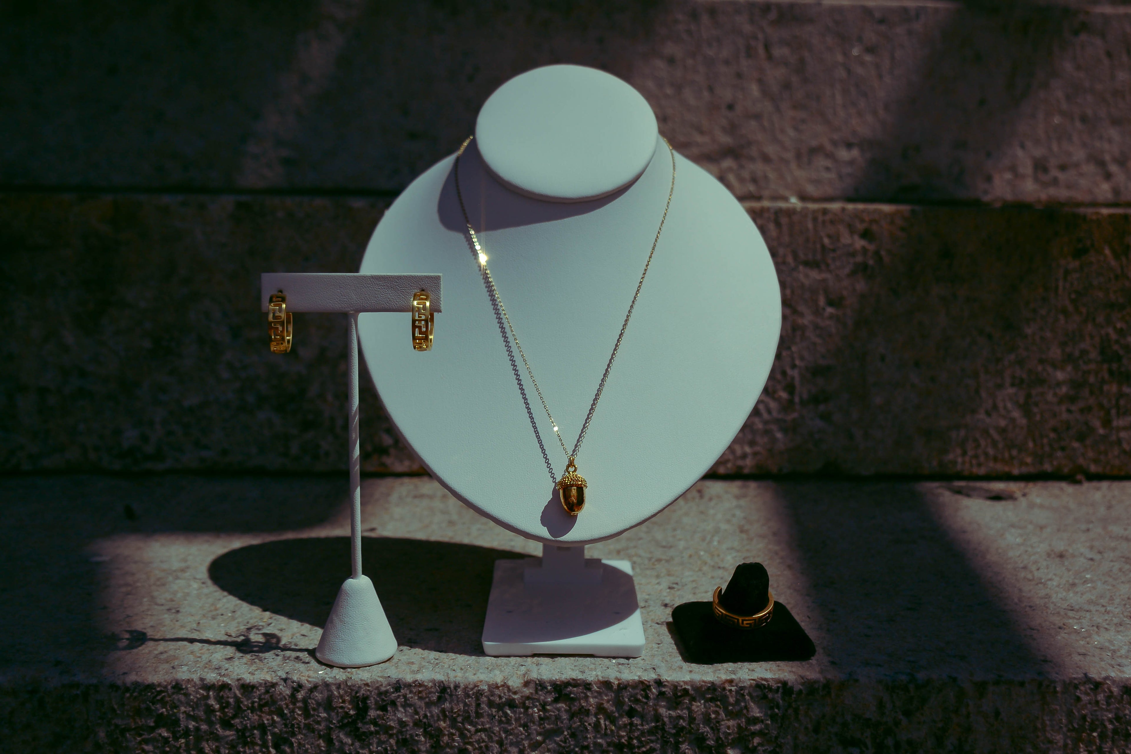 Gold dainty acorn necklace on white neck bust. Displayed alongside meander ring and earrings. On a granite rock background with creative purple lighting. 