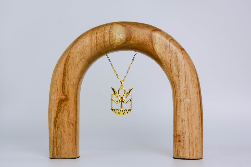 Golden ankh necklace with black detailing and center red gemstone. Necklace is suspended on a sleek wooden arch, on off-white background. 