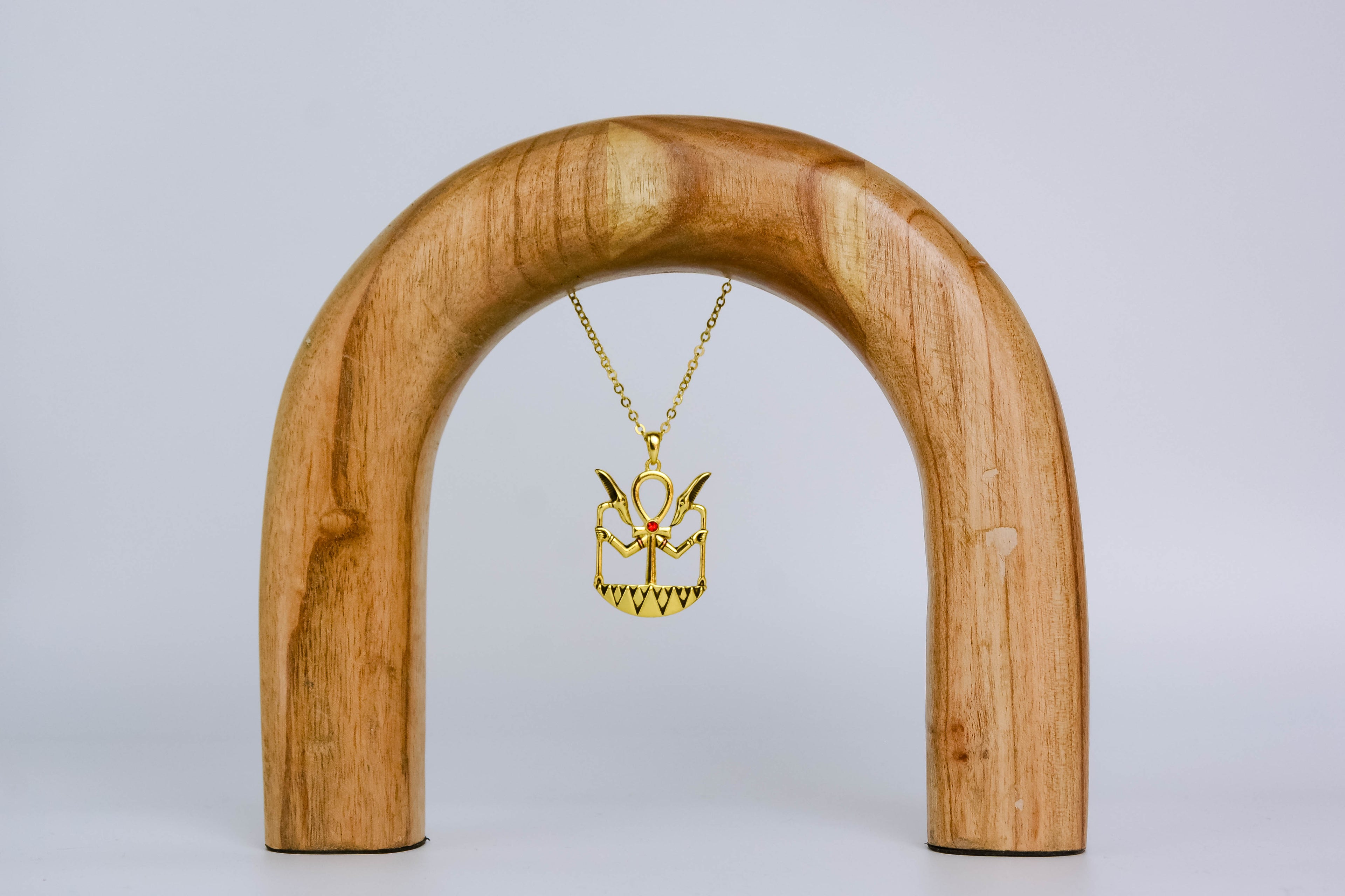 Golden ankh necklace with black detailing and center red gemstone. Necklace is suspended on a sleek wooden arch, on off-white background. 