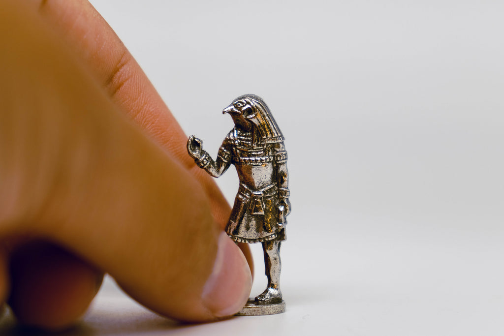 Horus figurine, silver with black detailing, on off-white background. Hand is picking up figuring for size comparison. 