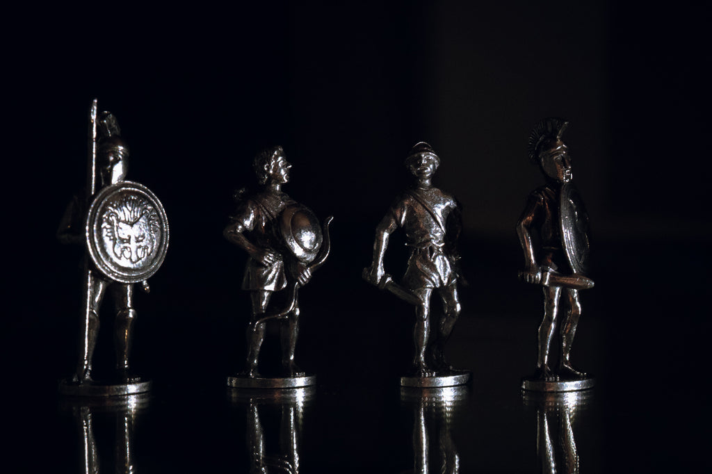 Four silver-colored metal Greek figurines. Reflected on the bottom and with various lights in background.