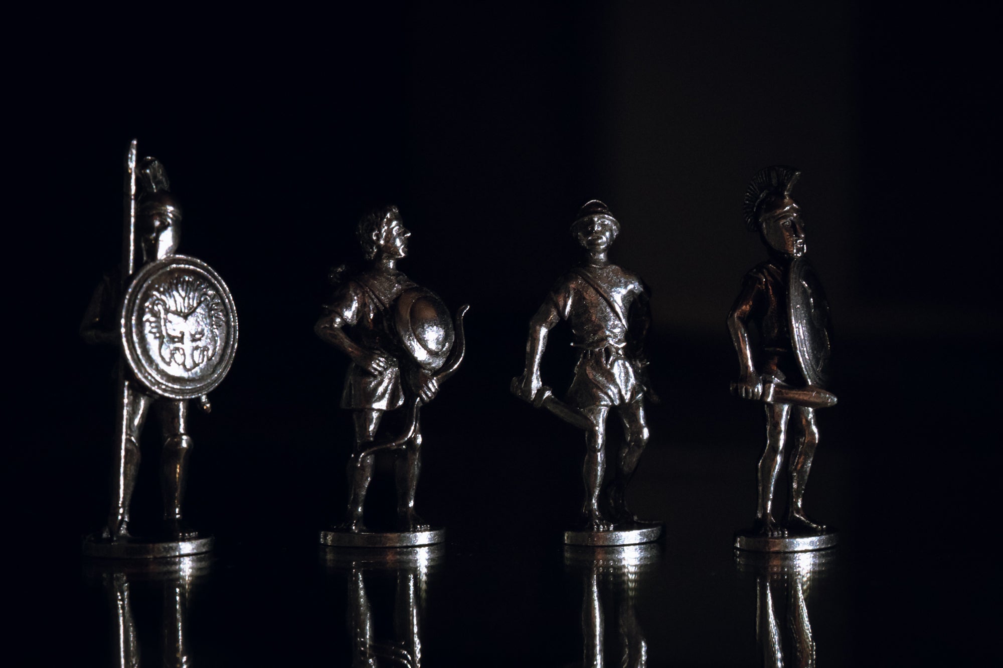 Four silver-colored metal Greek figurines. Reflected on the bottom and with various lights in background.