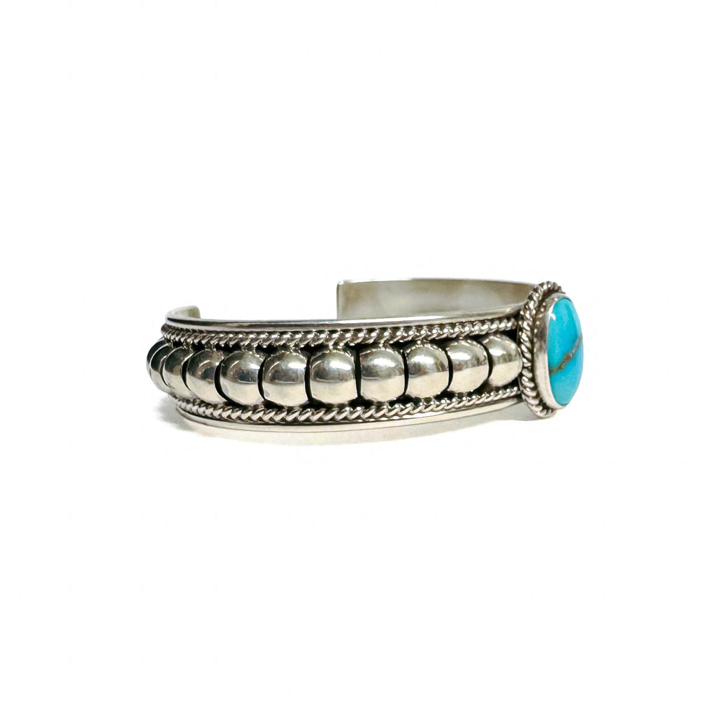 Side view of a silver bracelet featuring artistic accents and a turquoise stone at the center (facing right). Turquoise stone has a natural vein running through the center. Bracelet is on a white background.