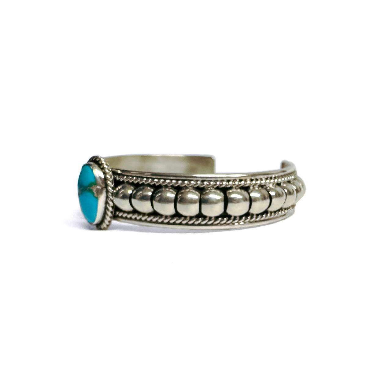 Side view of a silver bracelet featuring artistic accents and a turquoise stone at the center (facing left). Turquoise stone has a natural vein running through the center. Bracelet is on a white background.